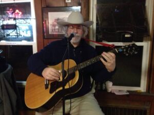Grey haired and bearded man wearing glasses and a grey cowboy hat is seating in front of a microphone and playing an acoustic guitar