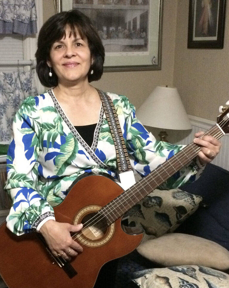 A women with chin length dark brown hair is holding an acoustic guitar. She is smiling and wearing a white blouse covered in blue and green tropical leaves.