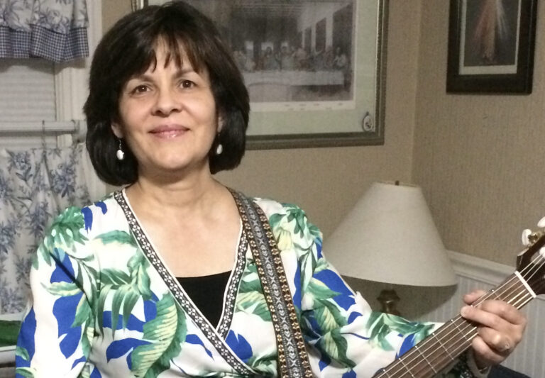 A women with chin length dark brown hair is holding an acoustic guitar. She is smiling and wearing a white blouse covered in blue and green tropical leaves.