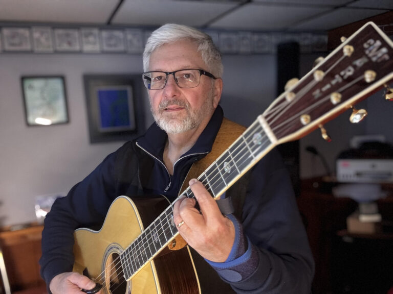 Man with grey hair and beard is holding an accostic guitar and looking at the camera