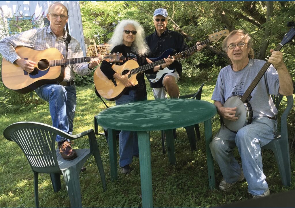 two men and a woman are holding guitars and standing behind a green table. A man holding a banjo is seated next to that table.