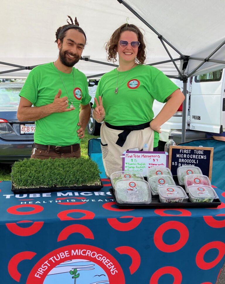 Smiling man and woman wearing bright green tshirts standing behind a table full of microgreens