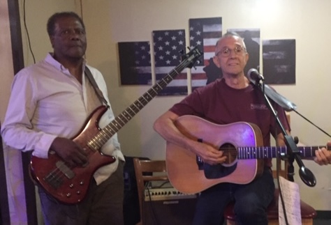 A Black man wearing a white shirt is holding an electric guitar and standing next to a White man in a maroon tshirt holding an acoustic guitar.