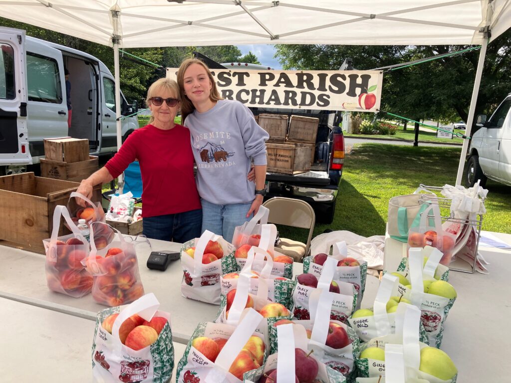 Two smiling women standing behind a table covered in tote bags full of peaches and red and yellow apples